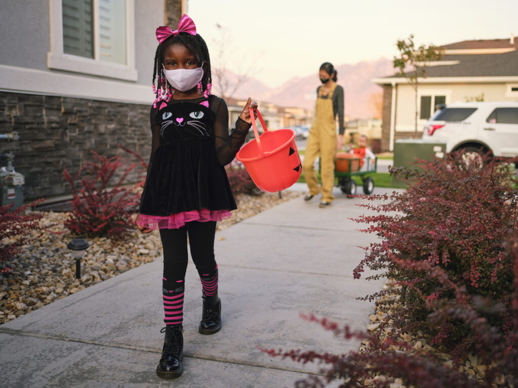 children trick or treating: African American girl in black cat costume in foreground, mom pulling kids in wagon in background