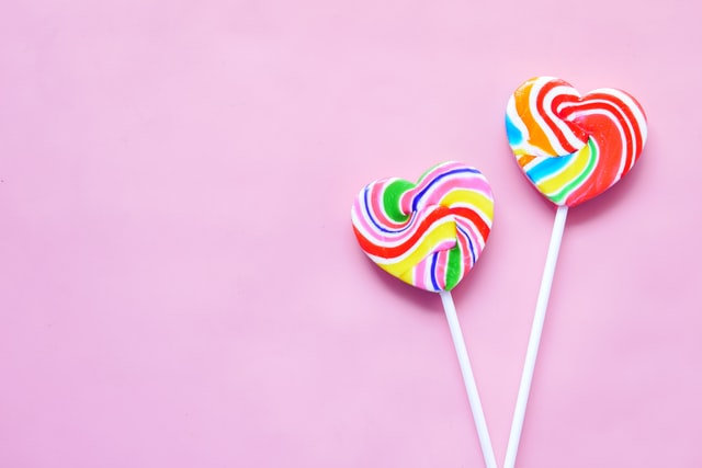 two heart-shaped rainbow swirl lollipops against pink background